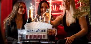 Three women celebrating at a nightclub table with a Grey Goose ice bucket holding champagne and vodka bottles, sparklers lit, surrounded by glasses and drinks."