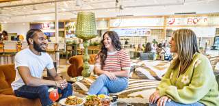 Three people enjoy expertly crafted food and beverages with smiles, gathered at a table in the brightly lit and beautifully decorated Politan Row food hall in Peachtree Corners.