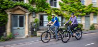 Two cyclists riding bikes through picturesque Charlbury, passing stone cottages covered with greenery