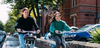 Two female students cycling past Queen's University on Belfast Bikes