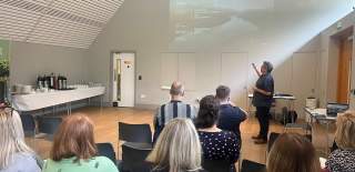 A group of people watch a presentation by a man, who gestures at a slide projection