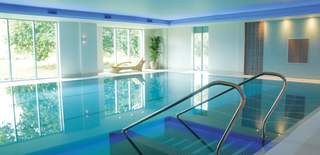 Sunlight pours through floor to ceiling windows over the still surface of a large indoor pool at De Vere Cotswold Water Park