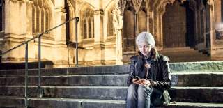 A woman sitting on the steps of St Mary Redcliffe Church wearing headphones listening to the Bristol audio tour