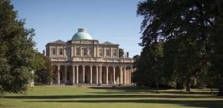 A sweeping lawn with large trees leads to the Grade I listed Regency Pittville Pump Room with green domed roof and large pillars