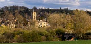 Wide countryside view showing Charlbury with stone houses and a church tower, surrounded by trees, fields, and a stone bridge under a partly cloudy sky.