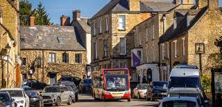 The centre of Charlbury with traditional stone-buildings lining both sides of the street, parked cars along both sides, a red bus driving through the centre, and warm sunlight highlighting the historic buildings.