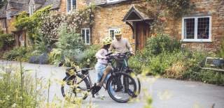 A couple have dismounted their bikes in front of a stunning cottage, with red roses climbing over it's porch, and are looking at their map