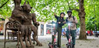 A couple on electric scooters pause to plan their way next to a large sculpture of animals sitting on a bench in Cheltenham, near the Cotswolds
