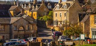 The centre of Blockley village with honey-coloured stone houses clustered along a quiet road, parked cars and people wandering on the village green in warm sunlight.