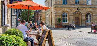 A group of people sit in the shade of an orange parasol outside a cafe, people sit on benches outside the adjacent town hall, whilst two ladies walk along shopping. Woodstock in the Cotswolds