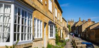 A couple wander down a sunlit village street in Blockley with honey-coloured stone cottages, large windows, and a narrow pavement lined with railings under a clear blue sky.