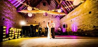 A newly married couple dance in the centre of a historic barn with fairy lights hanging from the wooden beamed ceiling at Cogges Manor Farm in the Cotswolds