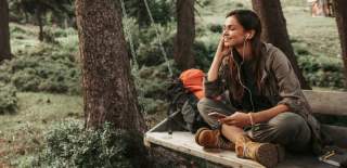Kvinne sitter på en benk i skogen med ørepropper og lytter til musikk