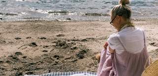 Kvinne som sitter på stranden på et badehåndkle med mat og drikke. Foto.
