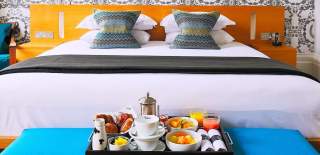 A breakfast tray filled with fresh fruit and pastries rests on a blue ottoman at the foot of a bed at Malmaison Cheltenham