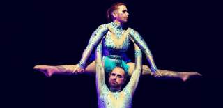 Two people wearing blue leotards in an acrobatic position. One person is holding another person in their air under their armpits.