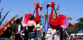 Two large flamingo puppets controlled by people inside them. There are three children looking at them with their backs to the camera.