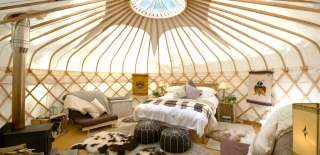 A circular skylight illuminates a cozy double bed, faux animal skin rugs and pouffes dotted in front of a wood burner inside Sapperton Yurt, at Westley Farm in the Cotswolds