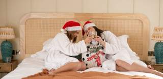 Two girls sitting on a hotel bed wearing santa hats holding glasses of prosecco