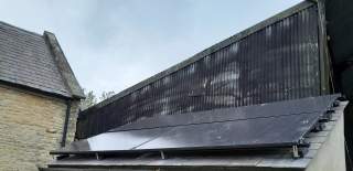 Modern building roof fitted with a row of solar panels, set beside a stone structure under an overcast sky.