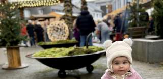 En liten jente i rosa vinterdress og lue med små dusker står på brosteinsplassen under julemarkedet i Lyngdal, med julelys, boder og mennesker i bakgrunnen.