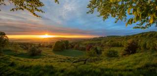 The sun rises over the horizon, illuminating the sky with a warm yellow glow over the lush green fields and trees surrounding the Cotswold Way