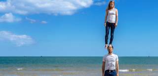 Two people on a beach wearing blue jeans and white tops. One person is standing on the head of another person.