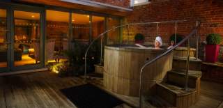 A lady relaxes at night in an outdoor wooden hot tub, surrounded by plants and subdued lighting at The Greenway Hotel & Spa in the Cotswolds