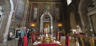 A couple with a tour guide stand in an impressive room with painted images on the ceiling and walls, large marble doorways and gold furniture at Blenheim Palace