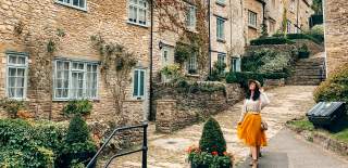 A woman wearing a hat smiles as she poses in front of stone cottages on a stepped hill in Tetbury