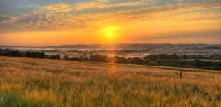The sun shines warm yellow through wispy clouds over open countryside, where mist lingers in the valleys