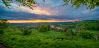 The sky glows with the start of sunrise over rolling green fields and trees viewed from the Cotswolds Way