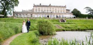 A bride and groom walk beside a lake with Cowley Manor in the background