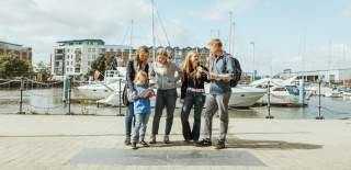 A group of people standing beside a stone fish carving on Hull’s Fish Trail near the marina, holding trail maps with boats and water in the background.
