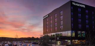 Photograph showing a Village Hotel at dusk, with the sun setting and creating a purple and orange-hued sky
