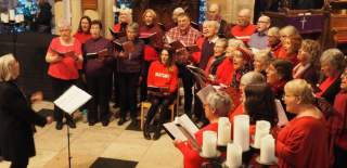 Beverley Community Choir at one of their carol concerts. The members are dressed in red jumpers and standing in a church. A woman in black leads at the front facing them.