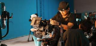 A production company in the filming process. Two men stand near a large film camera with lighting nearby.