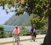 Two people wearing bike helmets pedal bicycles on the road near Mackinac Island boardwalk with water in the background