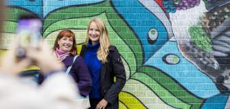 two women posing for a cell phone photo in front of a colorful mural of a bird