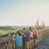 A wide, high-angle shot of a family of four looking out at the panoramic view from an elevated wooden boardwalk at Silver Dollar City in Branson, Missouri. On the left, a loop of an orange roller coaster track rises into the frame. In the center, a man in a purple shirt and khaki shorts stands with a young boy in a bright blue shirt to his left, and a young woman in a pink top and denim shorts to his right. Next to her stands another woman in a pink sleeveless shirt and shorts. All four have their backs to the camera, leaning against a rustic wooden railing to admire the vast expanse of rolling green Ozark hills stretching across the horizon under a clear sky. In the background on the right, the large, sweeping metal tracks and loops of the Wildfire roller coaster stand tall against the bright, sun-lit sky.