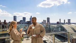 Couple in dressing gowns drinking coffee on Hotel Balcony overlooking Manchester