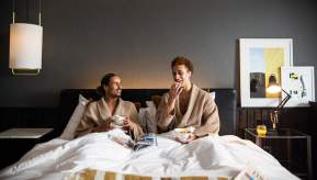 Couple in hotel bed in dressing gowns eating breakfast and drinking coffee
