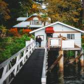 Two people stand on a narrow walkway leading to a small oceanfront house on stilts above the water, surrounded by trees and fall foliage.