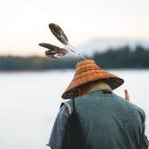 Person viewed from behind wearing a woven cedar hat decorated with feathers while standing near the shoreline.