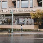 A person walks past The Coast Bastion Inn on a rainy day, with wet pavement and trees lining the street.