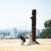 Three people stand at the base of a totem pole on Saysutshun with Nanaimo’s skyline visible across the harbour in the background.