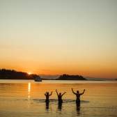 Three people standing in calm ocean water at sunset with arms raised.