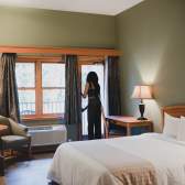 A cozy hotel room with a bed, desk, and seating area as a person opens a balcony door to let in natural light.