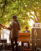 Booker T. Jones (on the piano) and his band members. The image is taken from the back of the stage and there is music equipment and speakers throughout the stage.