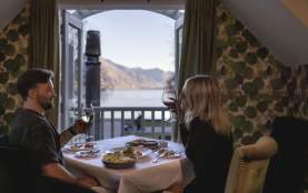 A cosy dinner setting with a couple enjoying wine and a delicious meal, overlooking stunning lake and mountain views through open balcony doors.